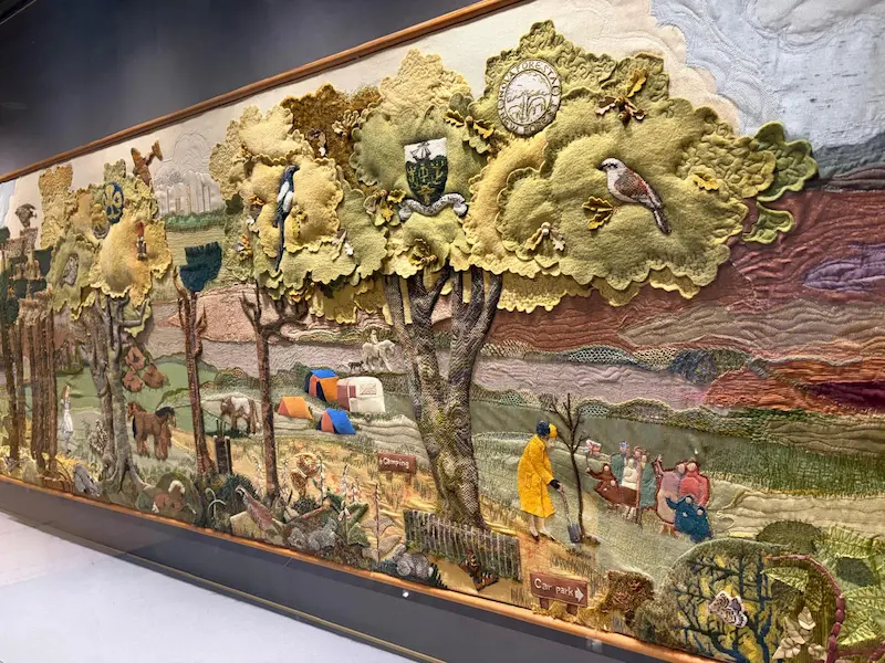 A large framed textile artwork depicting a pastoral English landscape, featuring embroidered and appliquéd scenes of trees, wildlife, people, and countryside activities.